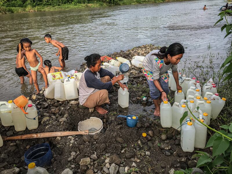 Ratna Jani (duduk) mengisi air bersih ke dalam jeriken sementara anaknya, Susan Jani, mengatur jeriken yang sudah terisi air bersih di pinggir sungai Desa Renggeang, Kecamatan Limboro, Kabupaten Polewali Mandar, Provinsi Sulawesi Barat, 11 Mei 2022. Ibu dan anak ini setiap sore mengambil air bersih di sungai untuk dijual kepada warga lainnya di desa seharga Rp1.000 per jeriken. Tampak di latar belakang anak-anak bermain di sungai. [Keisyah Aprilia/BenarNews]
