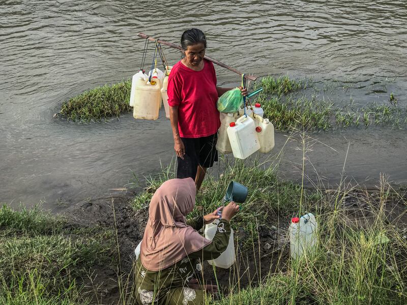 Nenek Samriati berdiri sambil memikul jeriken kosong menunggu keponakannya Indar Sari selesai mengisi air bersih ke dalam jeriken di pinggir sungai Desa Renggeang, Kecamatan Limboro, Kabupaten Polewali Mandar, Provinsi Sulawesi Barat, 11 Mei 2022. Hujan deras sejak dua hari terakhir di desa itu membuat air sungai meluap sehingga menyulitkan warga mendapatkan titik untuk penggalian mata air. Ketika satu mata air ditemukan, warga harus antre untuk bisa menggunakannya mengambil air bersih. [Keisyah Aprilia/BenarNews]