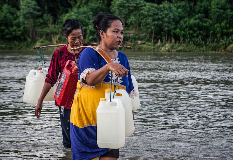Dua warga berjalan menyeberangi sungai sambil memikul jeriken berisi air bersih di sungai Desa Renggeang, Kecamatan Limboro, Kabupaten Polewali Mandar, Provinsi Sulawesi Barat, 11 Mei 2022. Sebagian warga yang bermukim di seberang sungai terpaksa harus menyeberangi sungai untuk mendapatkan air bersih. Air sungai tidak dalam sehingga tidak membahayakan mereka. Mayoritas warga di desa ini berjalan kaki untuk mengambil air bersih di sungai Renggeang. Meski jaraknya jauh antara tiga sampai empat kilo meter dari rumah mereka, warga tidak menjadikan itu beban. Menurut mereka ketimbang harus mengeluarkan uang untuk membeli air bersih di pasar mending mereka mengambilnya di sungai secara gratis. [Keisyah Aprilia/BenarNews]