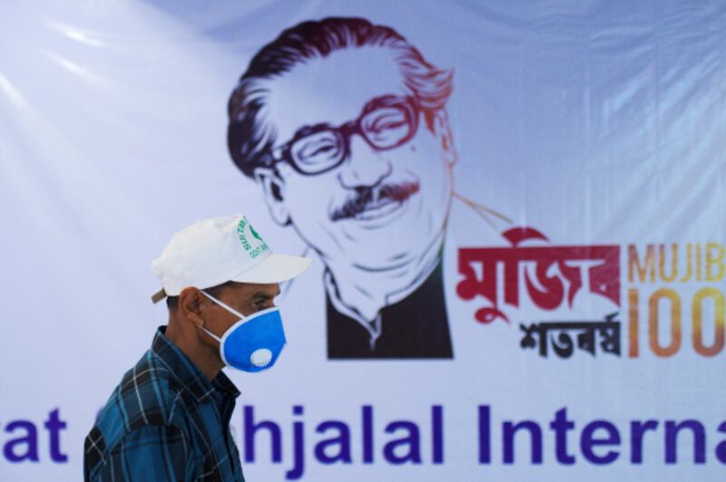 ঢাকার হযরত শাহ্জালাল আন্তর্জাতিক বিমানবন্দরে করোনাভাইরাস সংক্রমণ প্রতিরোধক হিসেবে মুখোশ পরে বঙ্গবন্ধুর জন্মশতবার্ষিকী অনুষ্ঠানের পোস্টারের সামনে দিয়ে হেঁটে যাচ্ছেন এক ব্যক্তি। বাংলাদেশে করোনাভাইরাস আক্রান্ত রোগী শনাক্ত হবার পর মুজিববর্ষের নির্ধারিত কর্মসূচি সংক্ষিপ্ত করেছে সরকার। ৮ মার্চ ২০২০।