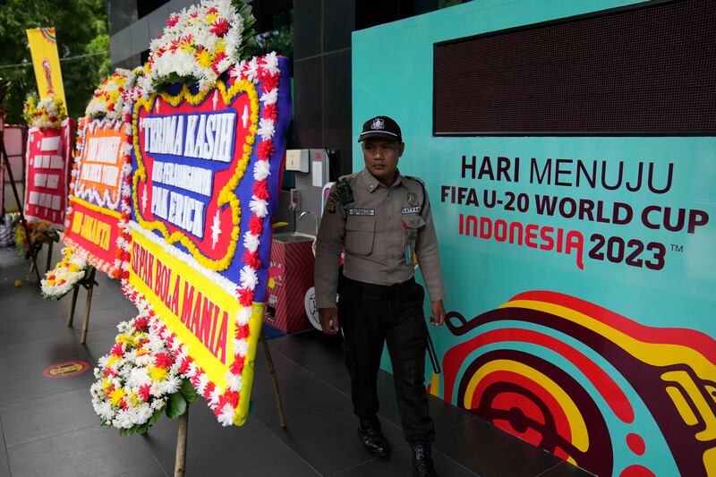 Seorang petugas berdiri di antara papan informasi pertandingan FIFA U-20 dan sejumlah karangan bunga tanda kekecewaan yang dikirim oleh para pendukung sepak bola di luar gedung PSSI di Jakarta, 30 Maret 2023. Sehari sebelumnya FIFA memituskan untuk membatalkan Indonesia sebagai tuan rumah Piala Dunia U-20, hanya delapan minggu sebelum dimulainya turnamen tersebut, menyusul gelombang penolakan sejumlah ormas Islam dan para politisi terkait partisipasi Israel dalam kejuaraan itu. [Dita Alangkara/AP Foto]